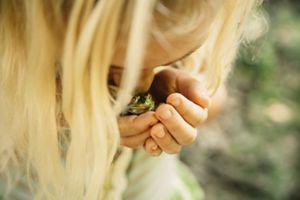 A young girl cups a frog in her hands and kisses its head.