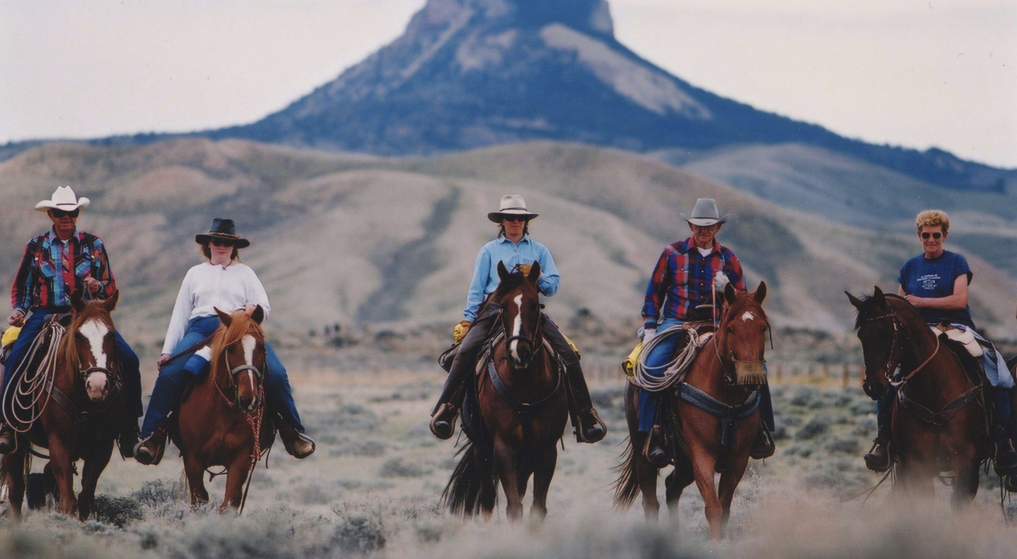 Five people on horseback riding towards the camera with a mountain in the background.