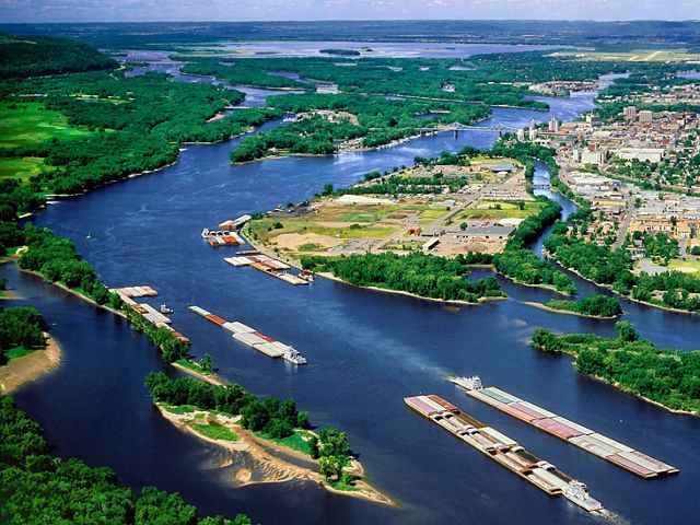 An aerial view of barges on the Mississippi river as it curves around populated and forested areas.