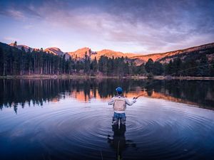 A person wearing waders stands knee-deep in a body of water surrounded by forested hills and casts a fly-fishing line.