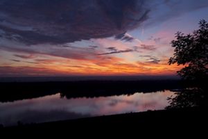 Sunrise illuminates wispy clouds with orange, pink, and purple hues over a still river in the foreground.