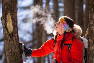 A smiling woman rests a gloved hand against a tree amidst a wintery forest.