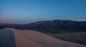 Group of people sitting atop a large sand dune in a desert setting with rugged mountains in the distance.
