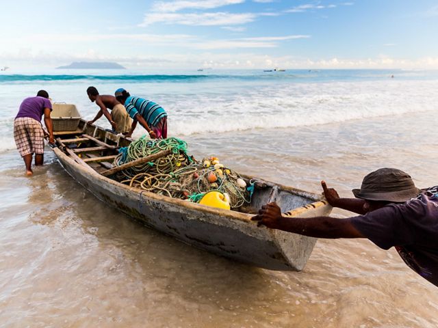 People pushing a small boat with fishing nets and gear through the waves into the sea.