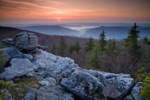 Aerial view looking out across a vast series of mountains, a valley shrouded in fog, and a setting sun under an orange sky.