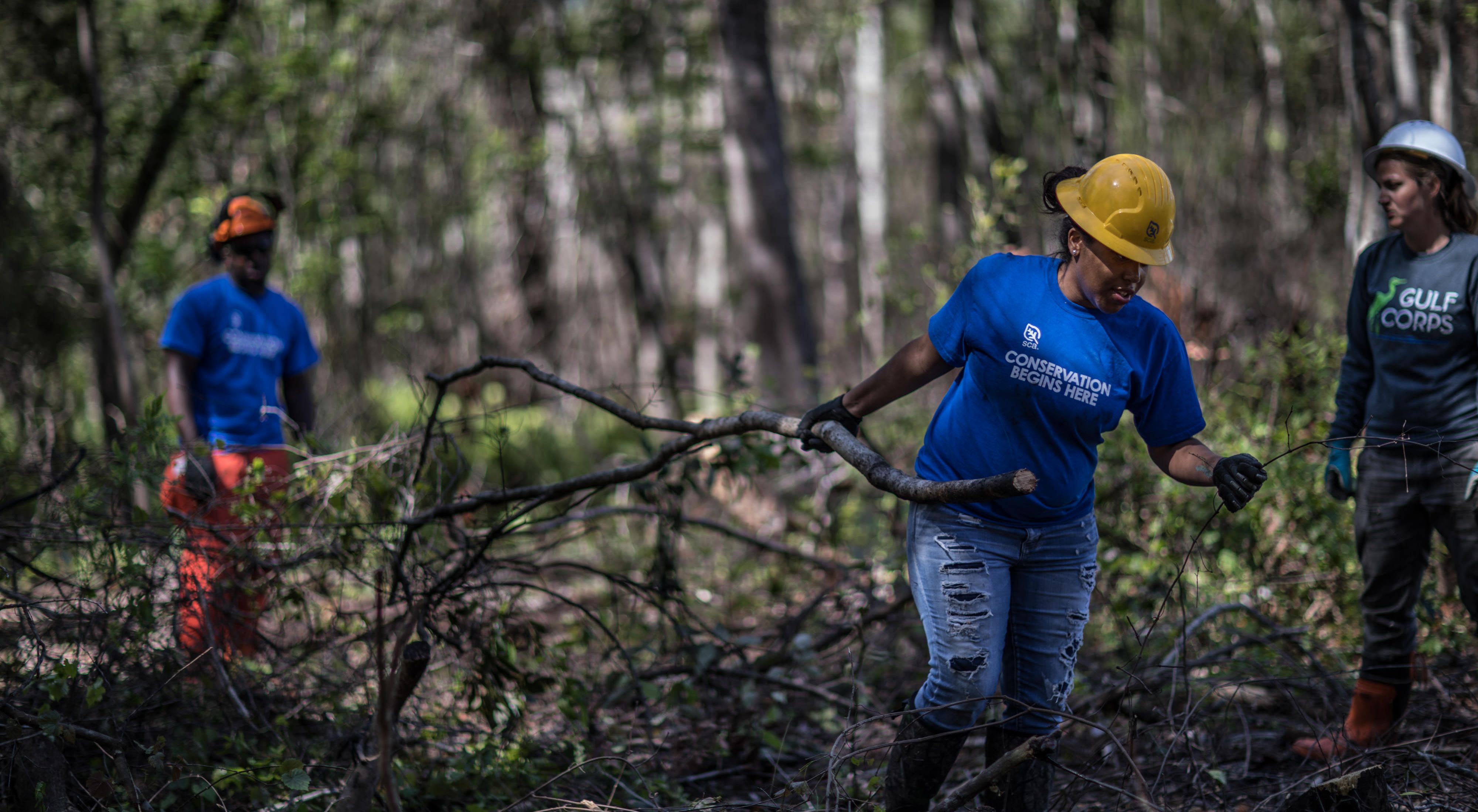 Three young people in hard hats, gloves and conservation corps shirts, clearing branches in a forest.
