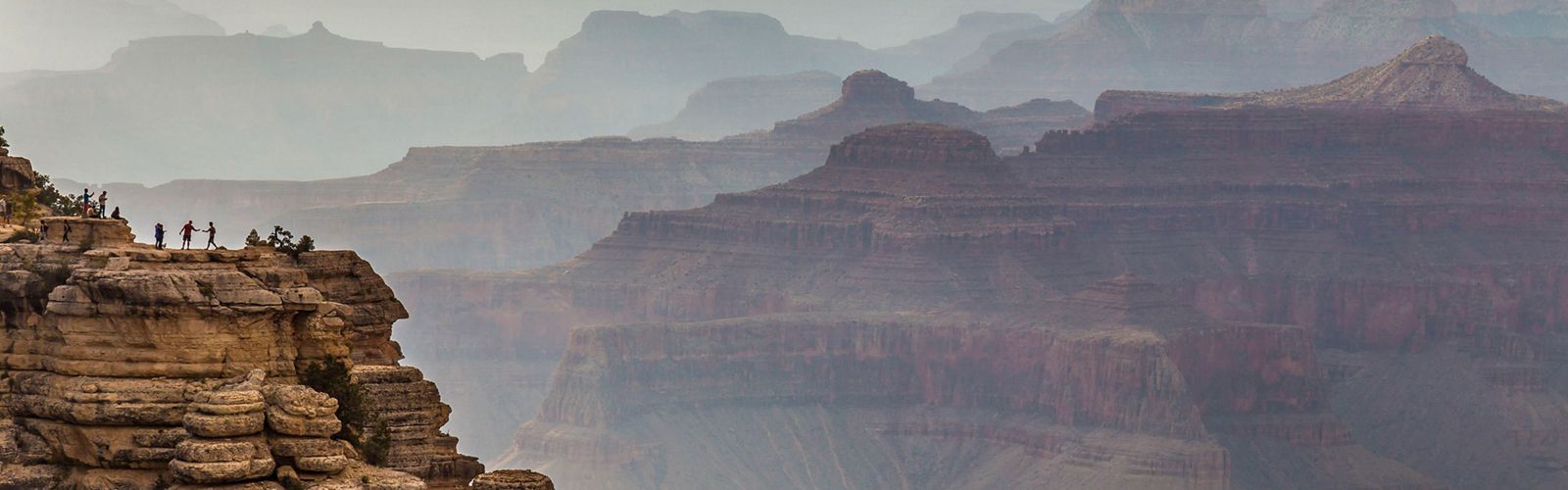 A few people stand atop a rock formation overlooking the Grand Canyon.