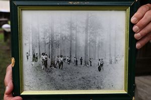 Man holding a black-and-white framed photo that shows African enslaved laborers working at a longleaf pine forest.