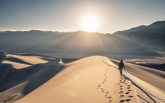 A lone figure walks along the crest of a tall sand dune, following a second set of footsteps. The sun hovers over the mountain range that lines the horizon.