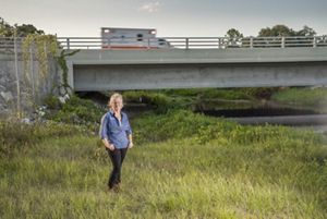 woman stands on grassy riverbank under a highway as an ambulance drives by