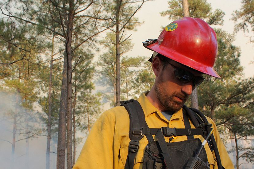 Sam Lindblom headshot. A man wearing fire gear looks down at a map as smoke rises behind him during a controlled burn.