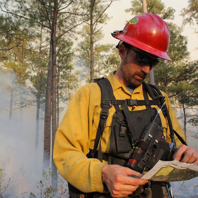 Sam Lindblom headshot. A man wearing fire gear looks down at a map as smoke rises behind him during a controlled burn.
