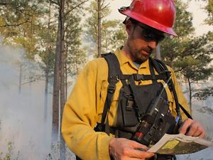 A man wearing yellow fire protective gear and a red helmet reviews a map during a controlled burn at Warm Springs Mountain. Thick smoke rises in the air behind him.