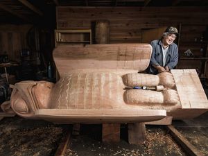 Within a carving shed, a man leans on a freshly carved totem pole of a sea wolf, made out of cedar wood