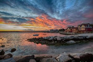 Sunset illuminates clouds with spectacular hues of orange, pink, and purple, with a tranquil waterfront and village in the foreground.