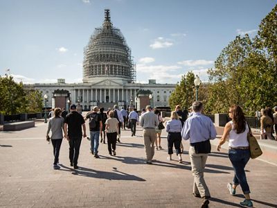 A large group of a dozen people walk alone the brick paved plaza towards the US Capitol building in Washington, DC. The building's dome is shrouded in scaffolding.