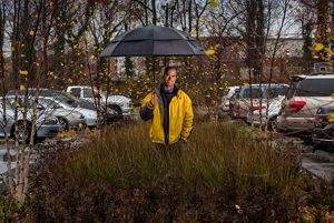 Kahlil Kettering stands holding an umbrella and wearing a yellow rain jacket in a grassy area in the parking lot of a church.