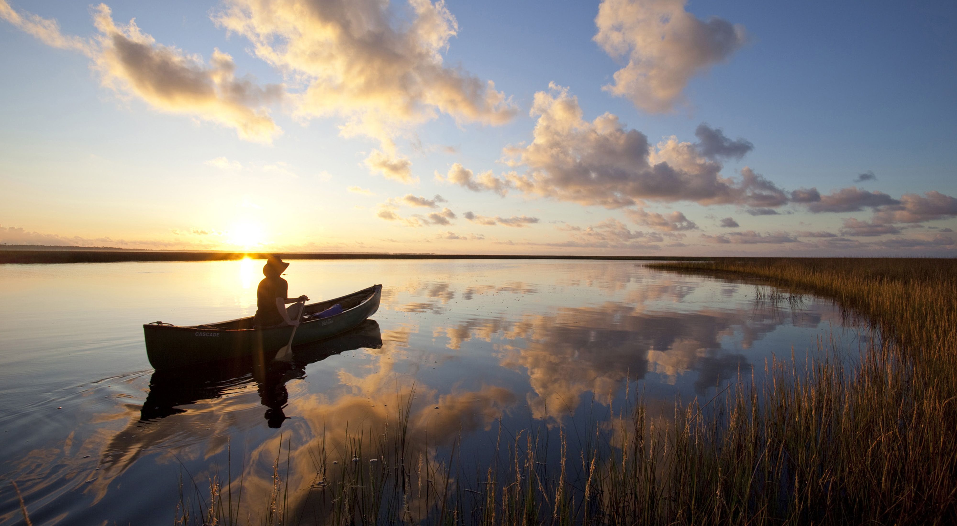 A person paddles a canoe at sunset on a marshy stream with fluffy clouds reflected in the surface of the water.