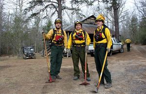 Three people wearing yellow protective fire gear pose together in front of a rustic cabin during a controlled burn on TNC's Warm Springs Mountain Preserve.