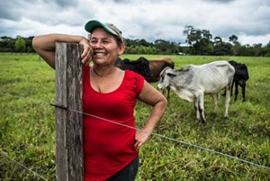 A woman in a baseball cap and red shirt is smiling and leaning against a fence. Her cows are standing behind her.