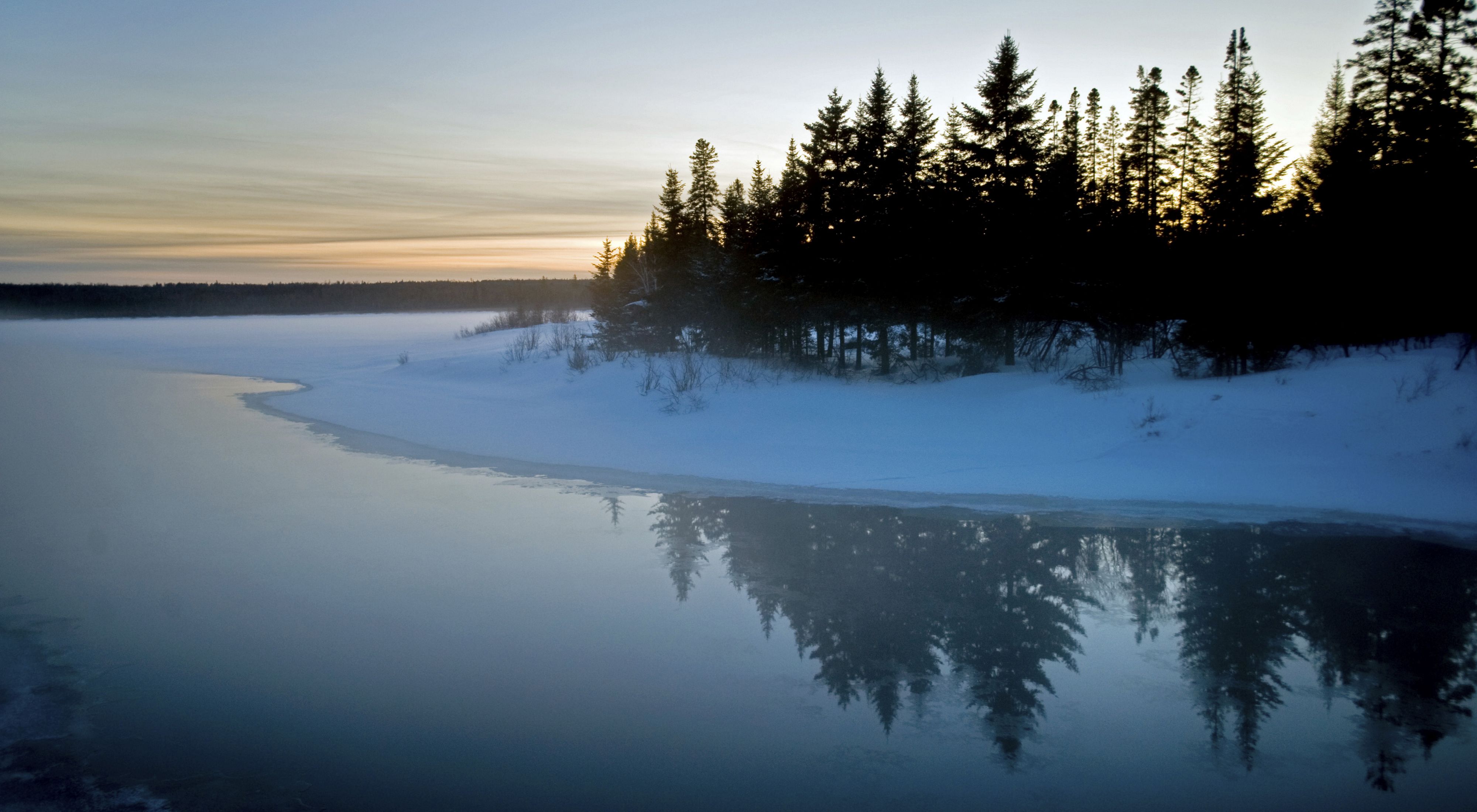 dusk on an open river bordered by snowy bank and evergreen trees