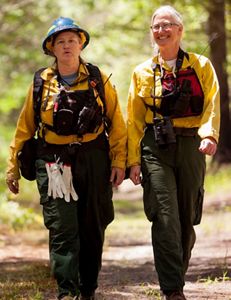Two women wearing yellow fire retardant gear walk together down a wide dirt path in a TNC preserve during a controlled burn. They both wear red vests holding walkie-talkies.