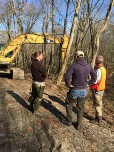 Three people stand near a yellow earth mover, a piece of heavy machinery used to cut breaches in the banks of the river behind them. The river is only barely visible through the trees.
