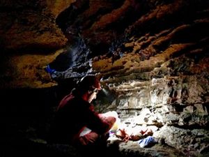 A man in an orange jumpsuit and hard hat crouches in a low cave preparing materials for a survey of the cave's bats. He uses his hat's headlamp to illuminate the pile of materials in front of him.