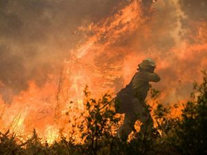 A firefighter dressed in fire-protective gear walks near a large wildfire.