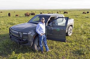 Man standing in front of electric pick-up truck in a prairie with buffalo behind him.