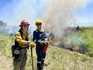 Two women wearing hard hats carry a drip torch while standing in front of a burning landscape.