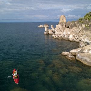 A person rows a red boat on a lake along a rocky shore.
