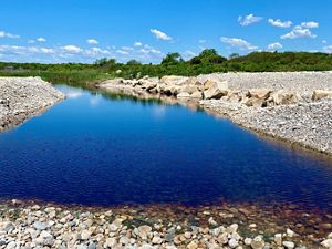A narrow channel of open water reflects the clear blue sky, with heaping piles of small stones forming both banks and green dune vegetation in the background.