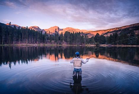 A person wearing waders stands knee-deep in a body of water surrounded by forested hills and casts a fly-fishing line.