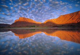 A canyon hillside and calm blue skies rflected in the water below.