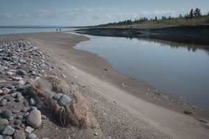 A sandy beach dotted with large pebbles. In the distance, two figures walk along the shore. 