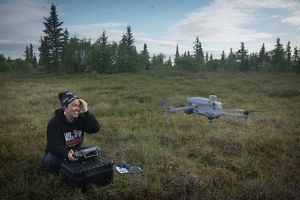Mary sits in the tundra and smiles as she uses a remote control to pilot a drone. 