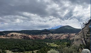 View looking out over green valleys, bare rock mesas and towering mountain peaks under low heavy clouds.
