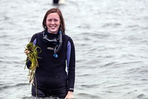 A smiling woman stands in thigh-deep water holding clumps of long, green eelgrass.