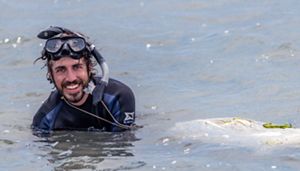 A smiling man in a wetsuit crouches in water that rises up to his shoulders. A mesh bag floats on the surface next to him.