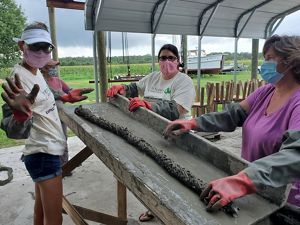 Three masked women stand around a tall table rolling burlap fabric in concrete to create oyster catching substrate for reef restoration.