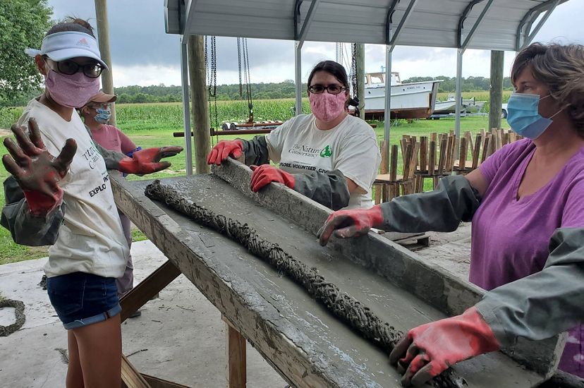 Three masked women stand around a tall table rolling burlap fabric in concrete to create oyster catching substrate for reef restoration.
