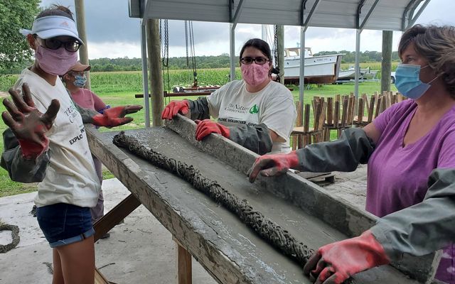 Three women stand around an elevated table using a concrete mixture to create a long, thin piece of oyster reef substrate.