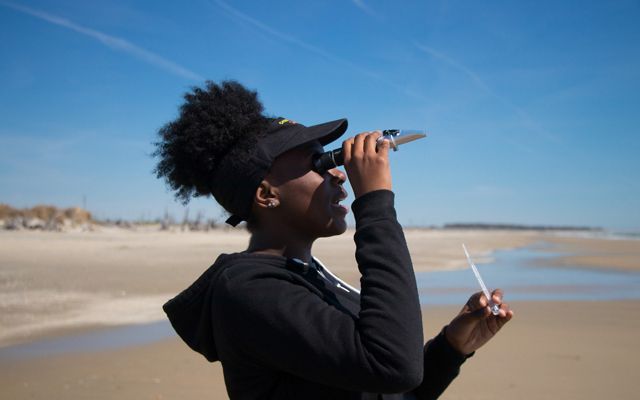 A girl holds a monocular to her eye while conducting science experiments on a beach.