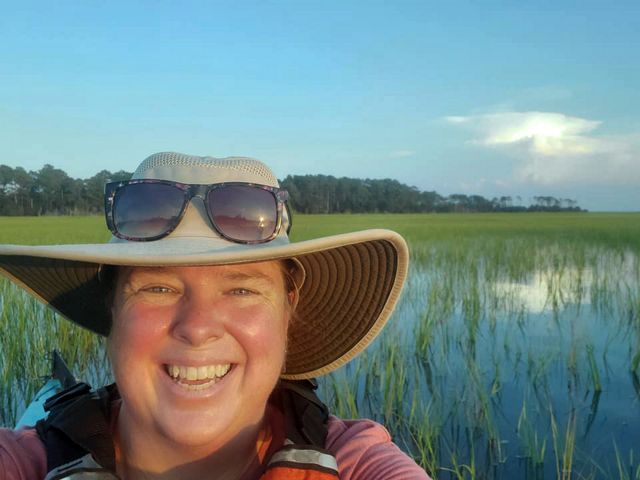Selfie portrait taken by a smiling woman sitting in a kayak. She is floating in a narrow coastal bay. Marsh grasses fill the water behind her.