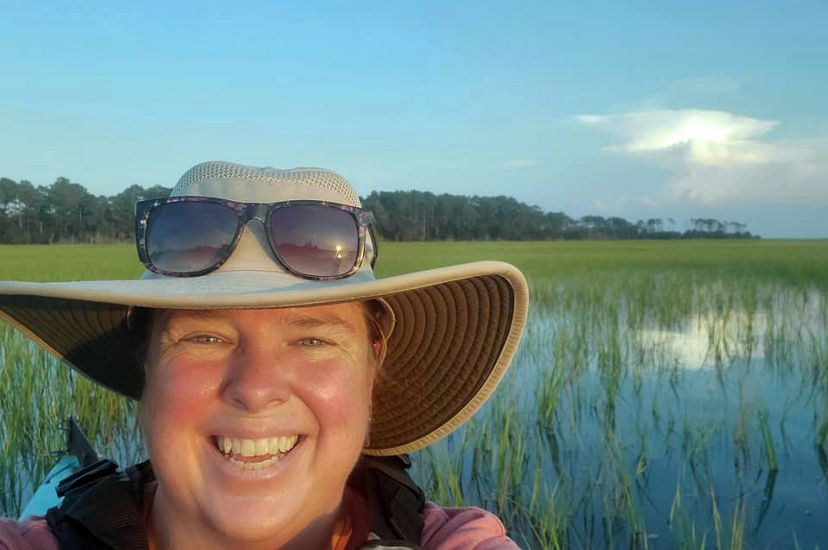Selfie portrait taken by a smiling woman sitting in a kayak. She is floating in a narrow coastal bay. Marsh grasses fill the water behind her.