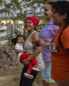 3 women stand mid-movement smiling, one holds a toddler in her arms that is laughing, fence and trees in background