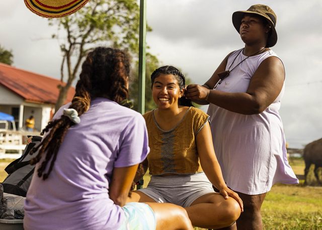 3 women gather: 2 sitting and talking and 1 braiding one of the first women's hair. A horse, barn and other ranch life elements in background