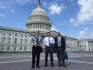Three men stand dressed in shirt and tie in front of the U.S. Capitol.