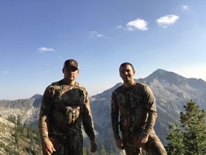 Two men in camouflage stand in front of a mountain ridge.